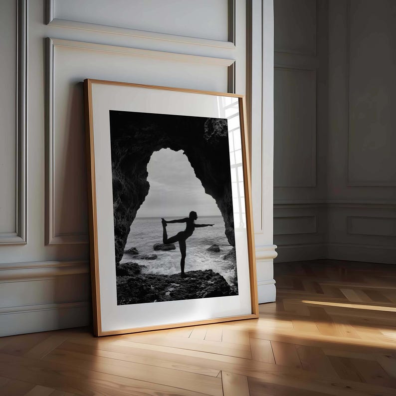 K&ouml;nnte beinhalten: Gerahmtes Schwarz-Wei&szlig;-Foto einer Person in einer Yoga-Pose, die auf Felsen am Meer steht. Das Bild befindet sich in einem hellbraunen Holzrahmen und lehnt an einer wei&szlig;en Wand.
