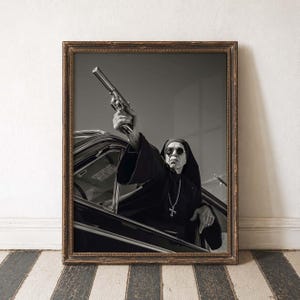 May include: Black and white framed print featuring a person dressed as a nun, wearing sunglasses, holding a handgun out of a car window. The print is in a wooden frame and is displayed against a white wall and a black and white striped floor.