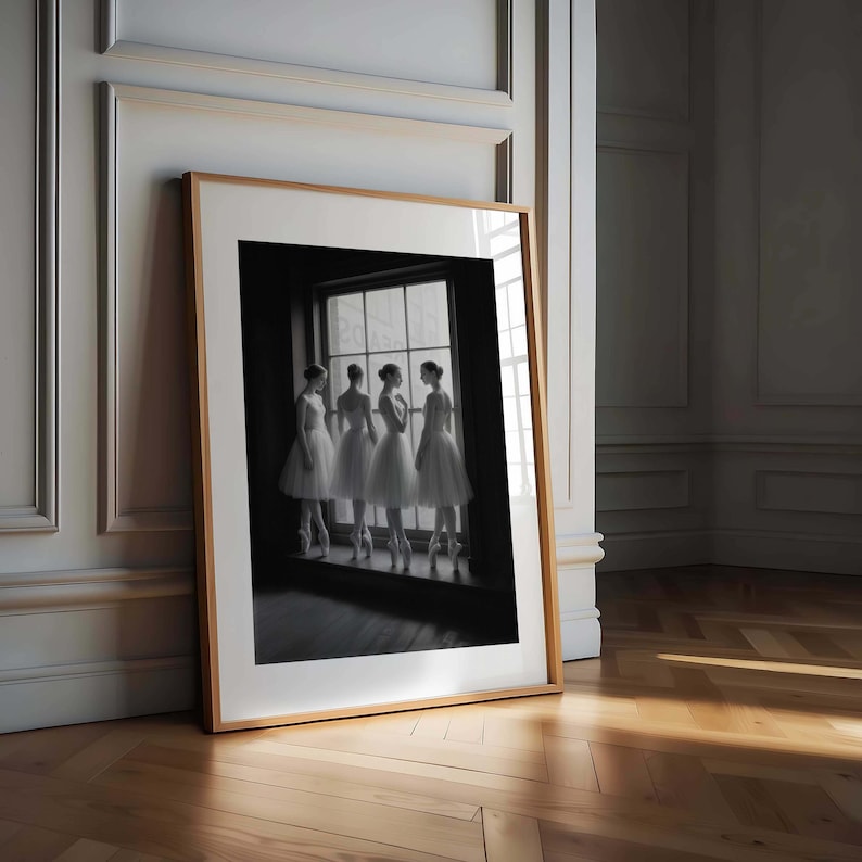 May include: A framed black and white photograph of four ballerinas in white tutus and pointe shoes, standing in a window. The photograph is in a light wood frame, leaning against a wall in a room with hardwood floors.