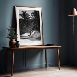 May include: Black and white framed print of a surfer riding a wave. The artwork features a surfer silhouetted against a crashing wave, with palm trees in the background. The frame is a light brown color, and the print is displayed on a wooden table.