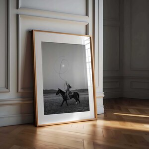 May include: A framed black and white photograph featuring a rider on horseback in a vast field, holding a lasso that forms a circular loop in the air against a bright sky. The artwork is encased in a light-toned wooden frame with a white mat.