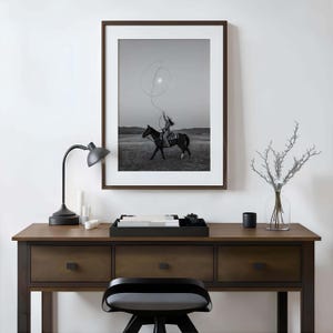 May include: A framed black and white wall art print depicting a rider on horseback swinging a lasso in an open field. The artwork is displayed above a dark wooden desk featuring a black desk lamp, a black tray with books, and a glass vase.