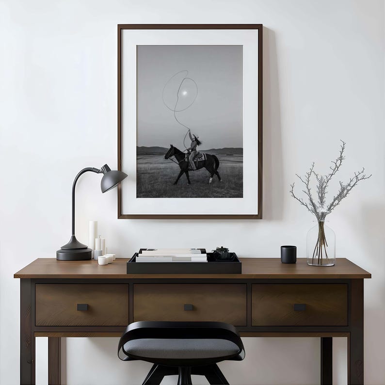 May include: A framed black and white wall art print depicting a rider on horseback swinging a lasso in an open field. The artwork is displayed above a dark wooden desk featuring a black desk lamp, a black tray with books, and a glass vase.