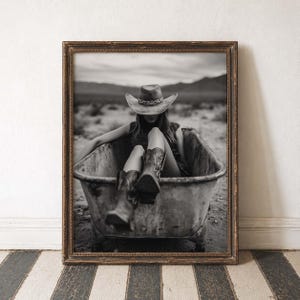 May include: Black and white framed photograph of a person wearing a cowboy hat and boots, sitting in a wheelbarrow. The image has a vintage aesthetic, with a desert landscape in the background.