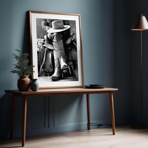 May include: Black and white framed print of a cowboy in a saddle, wearing a hat and cowboy boots. The boots have decorative stitching. The print is displayed on a wooden table with a plant and vase. The background is a dark blue wall.