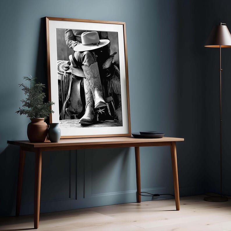 May include: Black and white framed print of a cowboy in a saddle, wearing a hat and cowboy boots. The boots have decorative stitching. The print is displayed on a wooden table with a plant and vase. The background is a dark blue wall.