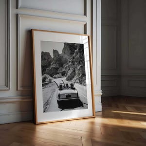 Puede incluir: Impresi&oacute;n enmarcada en blanco y negro de un coche descapotable en una carretera sinuosa a trav&eacute;s de monta&ntilde;as. Los pasajeros tienen los brazos levantados. La impresi&oacute;n se muestra contra una pared blanca y un suelo de madera.
