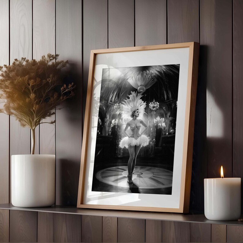 May include: Framed black and white print of a showgirl in a feathered headdress and costume, standing on a stage. The print is in a light wood frame, displayed on a shelf with a potted plant and a lit candle.