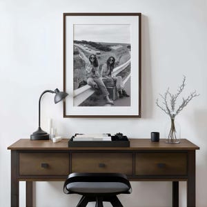 May include: Black and white framed photograph of two people sitting on a guardrail, one holding a sign that says "TAKE ME ANYWHERE". The photograph is displayed above a wooden desk with a black lamp and accessories.