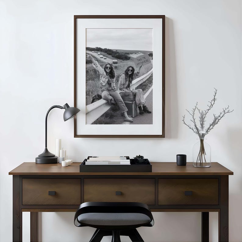 May include: Black and white framed photograph of two people sitting on a guardrail, one holding a sign that says "TAKE ME ANYWHERE". The photograph is displayed above a wooden desk with a black lamp and accessories.