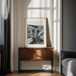 May include: A framed black and white photograph of two people hitchhiking on a highway, with one holding a sign. The photograph is displayed on a wooden cabinet with a black metal base, set against a neutral-toned wall.