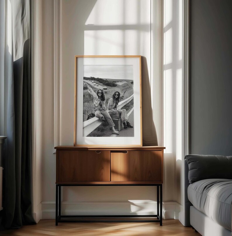May include: A framed black and white photograph of two people hitchhiking on a highway, with one holding a sign. The photograph is displayed on a wooden cabinet with a black metal base, set against a neutral-toned wall.