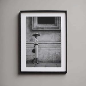 Könnte beinhalten: Schwarz-Weiß-Druck in einem Rahmen, der eine Person in einem Kleid und Hut zeigt, die auf einem Bürgersteig geht. Die Person trägt ein langärmeliges Kleid, einen breitkrempigen Hut und eine Handtasche. Der Hintergrund ist eine Steinmauer mit einem Fenster.