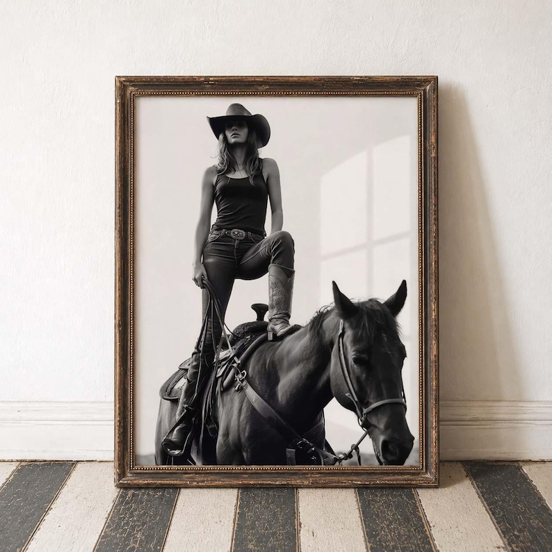 May include: Black and white framed print of a person in a cowboy hat and boots, standing on a horse. The print has a vintage aesthetic, with a dark wooden frame and a monochrome image.