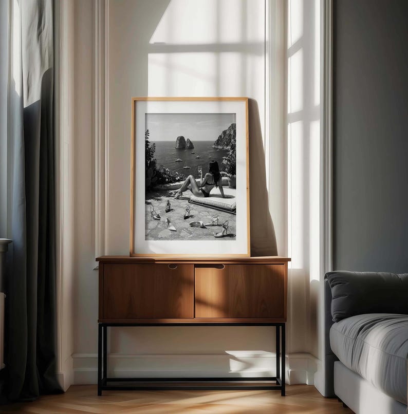 May include: A framed black and white wall art print featuring a person in a bikini lounging on a stone terrace overlooking the sea with large rock formations in the distance. Several pairs of high-heeled sandals are scattered on the ground.