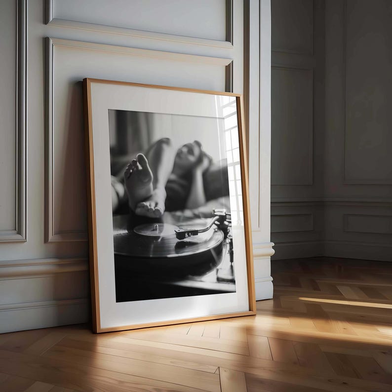 K&ouml;nnte beinhalten: Schwarz-Wei&szlig;-Druck in einem Rahmen, der eine Person zeigt, die sich entspannt und die F&uuml;&szlig;e auf einem Plattenspieler hat. Das Bild befindet sich in einem Holzrahmen, der an einer Wand lehnt. Der Raum hat ein klassisches, elegantes Design.
