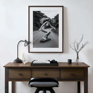 Peut inclure: Impression encadr&eacute;e en noir et blanc d'une personne faisant du skateboard sur une route. L'image est dans un cadre marron fonc&eacute;, au-dessus d'un bureau en bois. Une lampe de bureau noire, des bougies et un plateau d&eacute;coratif sont sur le bureau.