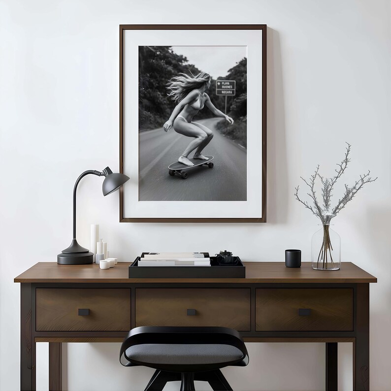 Peut inclure: Impression encadr&eacute;e en noir et blanc d'une personne faisant du skateboard sur une route. L'image est dans un cadre marron fonc&eacute;, au-dessus d'un bureau en bois. Une lampe de bureau noire, des bougies et un plateau d&eacute;coratif sont sur le bureau.