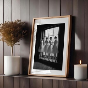 May include: Black and white framed print featuring four ballerinas in white tutus and pointe shoes, standing by a window. The image is set in a room with a wooden wall, a plant in a white pot, and a lit candle.