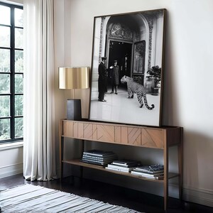 Op de afbeelding: Een zwart-witfoto van twee mannen in formele kleding die voor een gebouw staan met een luipaard dat naar hen toe loopt. De foto is ingelijst in een gouden lijst en hangt aan een witte muur boven een houten consoletafel.
