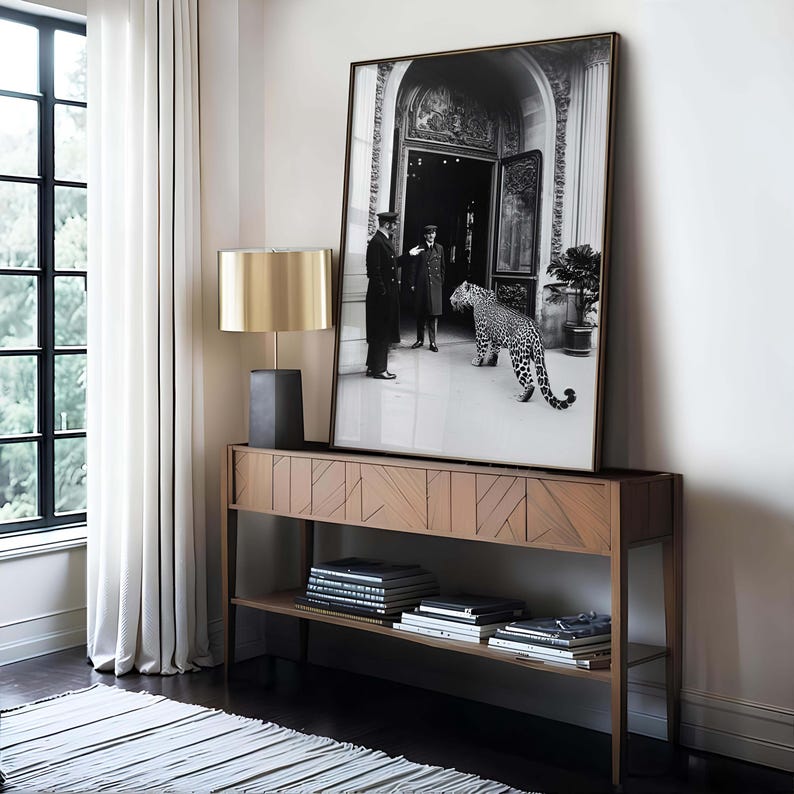 Op de afbeelding: Een zwart-witfoto van twee mannen in formele kleding die voor een gebouw staan met een luipaard dat naar hen toe loopt. De foto is ingelijst in een gouden lijst en hangt aan een witte muur boven een houten consoletafel.