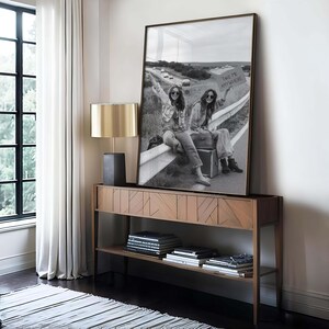 May include: A framed black and white photograph of two people hitchhiking, holding a sign that reads "TAKE ME ANYWHERE." The photograph is displayed on a wooden console table with a gold lamp. The room has a window with white curtains and a striped rug.
