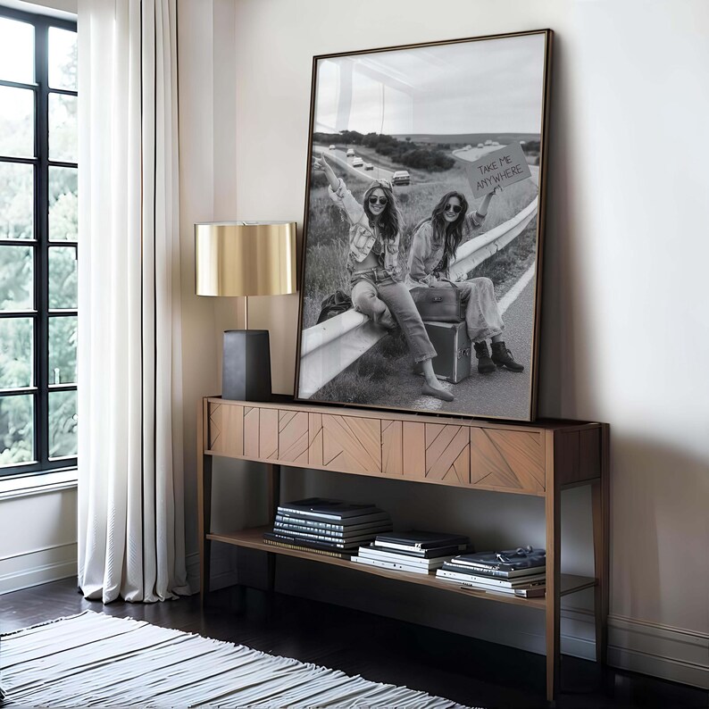 May include: A framed black and white photograph of two people hitchhiking, holding a sign that reads "TAKE ME ANYWHERE." The photograph is displayed on a wooden console table with a gold lamp. The room has a window with white curtains and a striped rug.