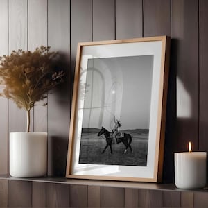 May include: A framed black and white photograph depicting a person riding a horse in an open field while swinging a lasso. The print is housed in a light-colored wooden frame, positioned between a white potted plant and a white pillar candle.