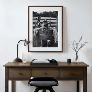 May include: Monochrome framed print of a person in a cowboy hat and boots, seated in a metal tub. The print is on a wooden desk with a black lamp, candles, and a decorative tray.