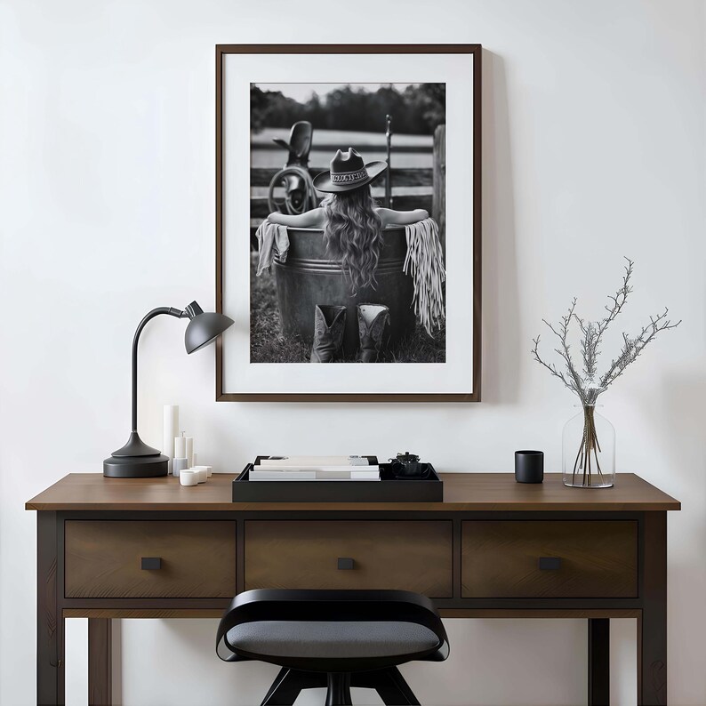 May include: Monochrome framed print of a person in a cowboy hat and boots, seated in a metal tub. The print is on a wooden desk with a black lamp, candles, and a decorative tray.