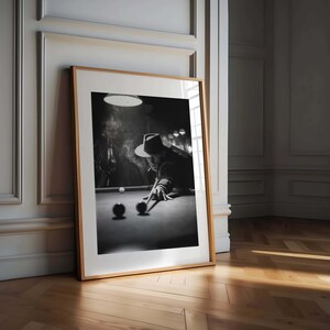 May include: Monochrome framed print depicting a person playing snooker, smoking a cigarette. The person wears a hat and leans over the snooker table, aiming with a cue. The print is in a wooden frame, leaning against a wall.