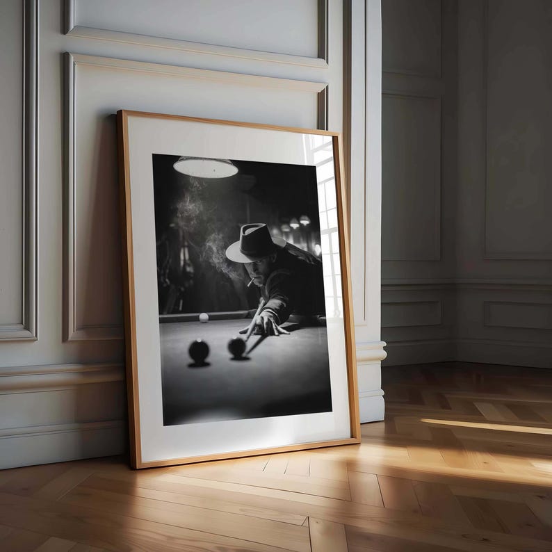 May include: Monochrome framed print depicting a person playing snooker, smoking a cigarette. The person wears a hat and leans over the snooker table, aiming with a cue. The print is in a wooden frame, leaning against a wall.