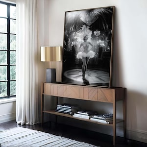 May include: Black and white framed print of a showgirl in a feathered headdress and tutu, standing on a stage. The artwork is displayed on a wooden console table with a gold lamp.