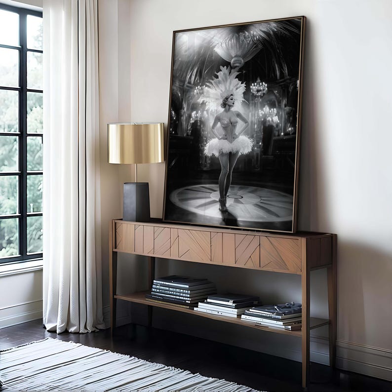 May include: Black and white framed print of a showgirl in a feathered headdress and tutu, standing on a stage. The artwork is displayed on a wooden console table with a gold lamp.