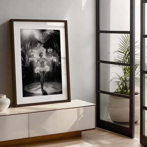 May include: Black and white framed print of a woman in a feathered costume, standing on a stage. The print is in a dark brown frame and is displayed on a white cabinet. The background shows a chandelier and other decorative elements.