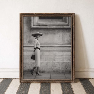 Puede incluir: Impresión enmarcada en blanco y negro de una mujer con vestido y sombrero, caminando por una acera. La mujer lleva un vestido claro, un sombrero de ala ancha y un bolso negro. El fondo presenta una pared de piedra.