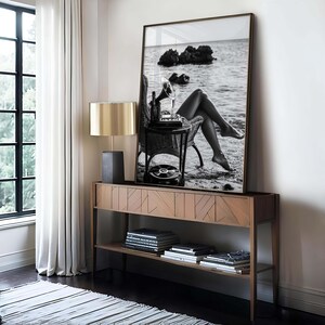 Pode incluir: Fotografia a preto e branco emoldurada de uma mulher sentada numa praia com uma gira-discos vintage. A fotografia &eacute; exibida acima de uma consola de madeira com uma l&acirc;mpada e livros.