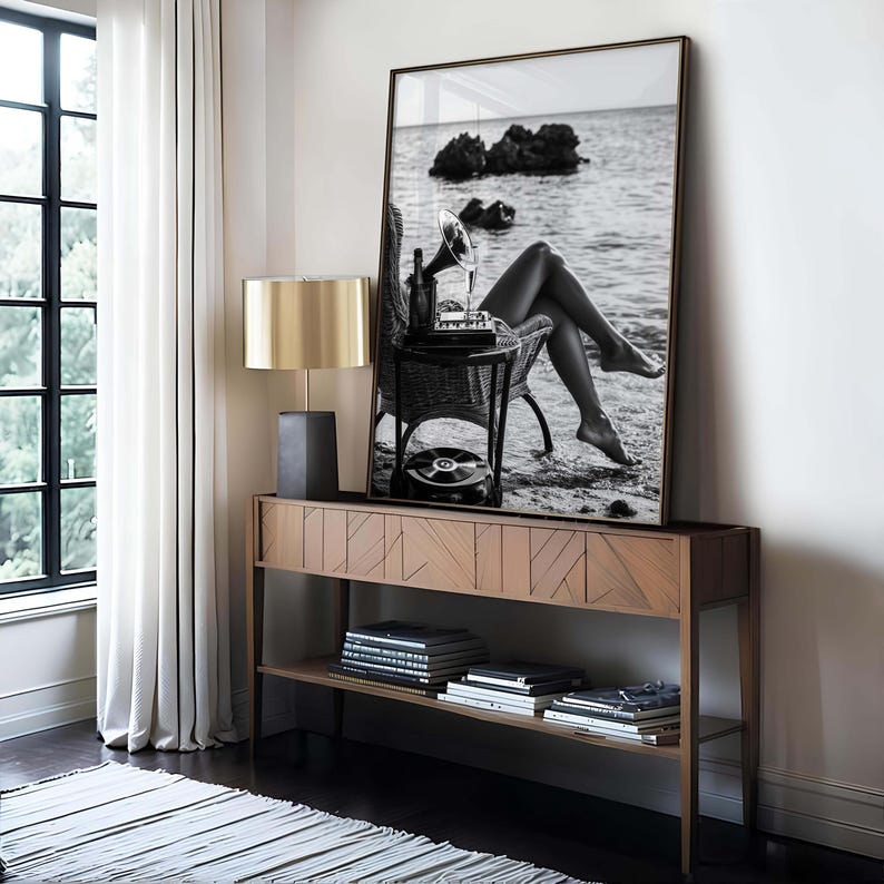 Pode incluir: Fotografia a preto e branco emoldurada de uma mulher sentada numa praia com uma gira-discos vintage. A fotografia &eacute; exibida acima de uma consola de madeira com uma l&acirc;mpada e livros.