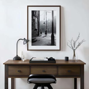 May include: Black and white photograph of two children playing in a narrow street. The photo is framed in a dark wood frame and is hanging on a white wall above a wooden desk with drawers.