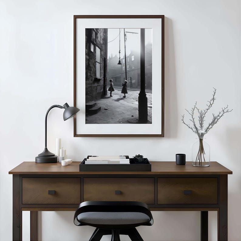 May include: Black and white photograph of two children playing in a narrow street. The photo is framed in a dark wood frame and is hanging on a white wall above a wooden desk with drawers.