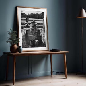 May include: Framed monochrome photograph of a person wearing a cowboy hat and boots, sitting in a metal tub. The photograph is displayed on a wooden table with a small plant and vase.