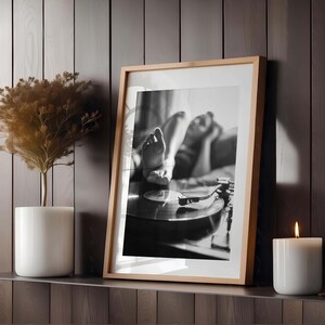 K&ouml;nnte beinhalten: Gerahmte Schwarz-Wei&szlig;-Fotografie einer Person, die sich in der N&auml;he eines Plattenspielers entspannt. Das Foto befindet sich in einem hellen Holzrahmen. Eine wei&szlig;e Kerze und eine Topfpflanze stehen auf einem Regal neben dem Foto.