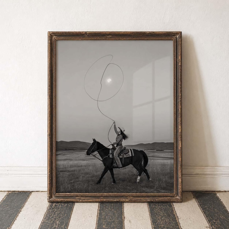 May include: A black and white framed wall art print depicting a person riding a horse in a vast field. The rider is swinging a lasso in the air against a bright sun. The scene features rolling hills in the distance, captured in a monochromatic style.