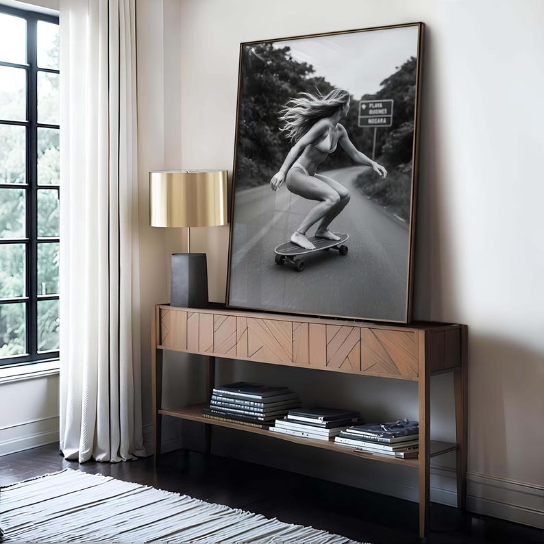 Peut inclure: Impression en noir et blanc d'une personne faisant du skateboard sur une route. L'image est affich&eacute;e sur une console en bois avec une lampe dor&eacute;e. La pi&egrave;ce a une fen&ecirc;tre avec des rideaux blancs et un tapis ray&eacute;.