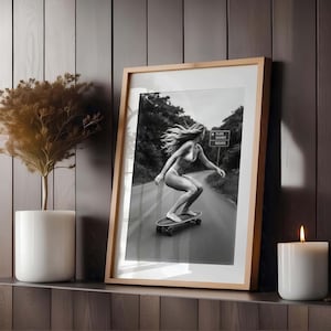Peut inclure: Photographie encadr&eacute;e en noir et blanc d'une personne faisant du skateboard sur une route. La personne porte un bikini et a de longs cheveux flottants. La photographie est dans un cadre en bois clair et est expos&eacute;e sur une &eacute;tag&egrave;re avec une bougie et une plante.
