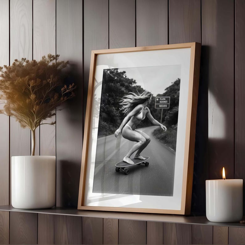 Peut inclure: Photographie encadr&eacute;e en noir et blanc d'une personne faisant du skateboard sur une route. La personne porte un bikini et a de longs cheveux flottants. La photographie est dans un cadre en bois clair et est expos&eacute;e sur une &eacute;tag&egrave;re avec une bougie et une plante.