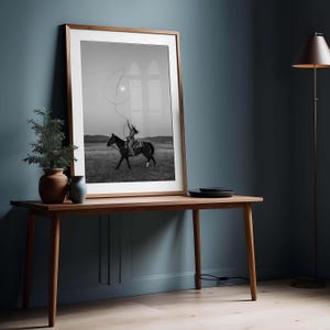 May include: A framed black and white wall art print depicting a rider on horseback swinging a lasso in an open field. The print is housed in a thin wooden frame with a white mat, resting on a wooden table alongside two small ceramic vases.