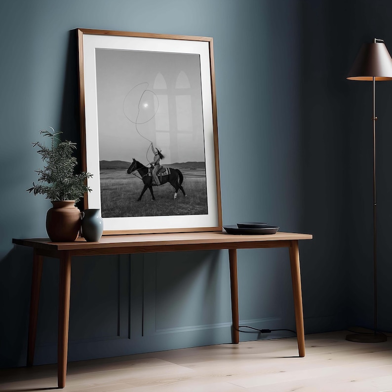 May include: A framed black and white wall art print depicting a rider on horseback swinging a lasso in an open field. The print is housed in a thin wooden frame with a white mat, resting on a wooden table alongside two small ceramic vases.