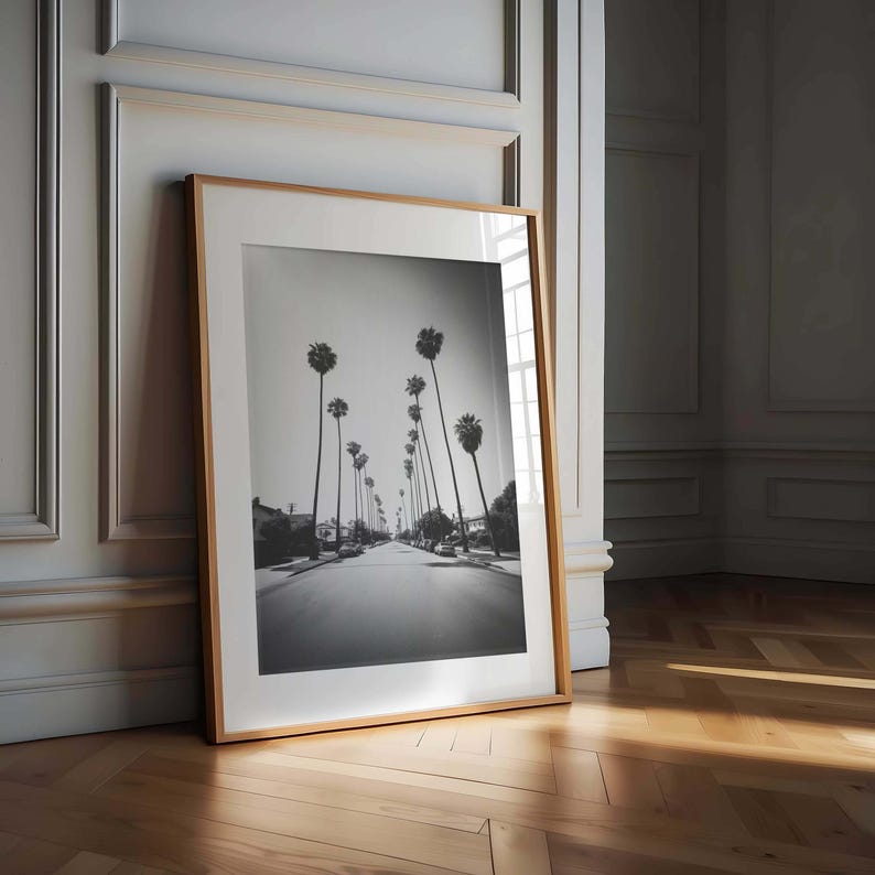 May include: Black and white framed print of a street lined with tall palm trees. The photograph is in a vertical orientation and is displayed in a light wood frame, leaning against a wall. The image captures a sunny day.
