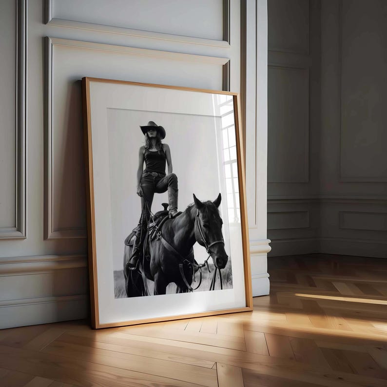 May include: Black and white framed print of a person in a cowboy hat and jeans standing on a horse. The print is leaning against a wall with a wooden frame and a light-colored mat.